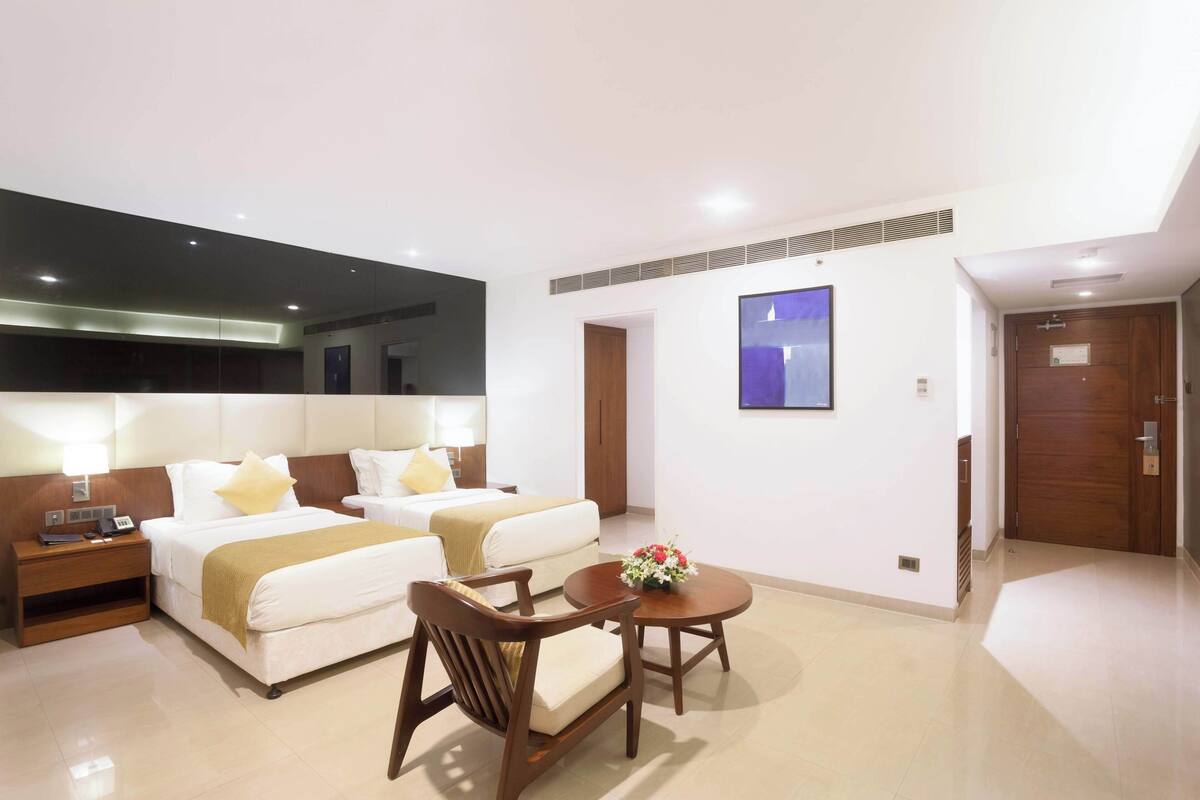 A hotel room with two beds, a desk, a chair, a small table with flowers, and a TV on the wall.