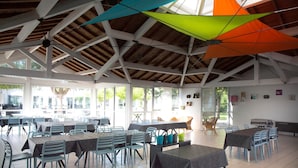 A spacious indoor dining area with multiple tables and chairs, a high ceiling with wooden beams, and large windows allowing natural light.