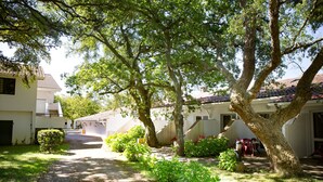 A residential area with a paved driveway leading to a house with a tiled roof. There are trees and shrubs in the foreground, and a small garden area with a bench.