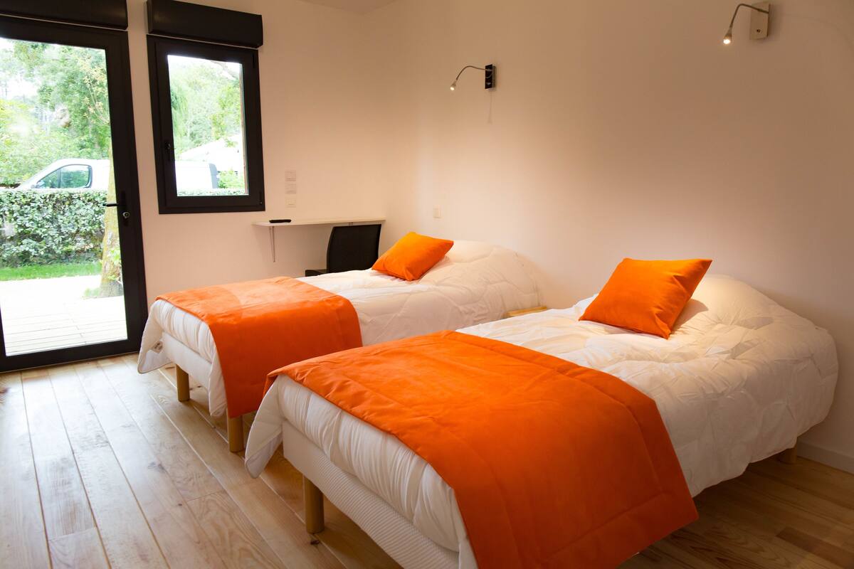 A room with two beds, each with an orange blanket, a desk, and a window.