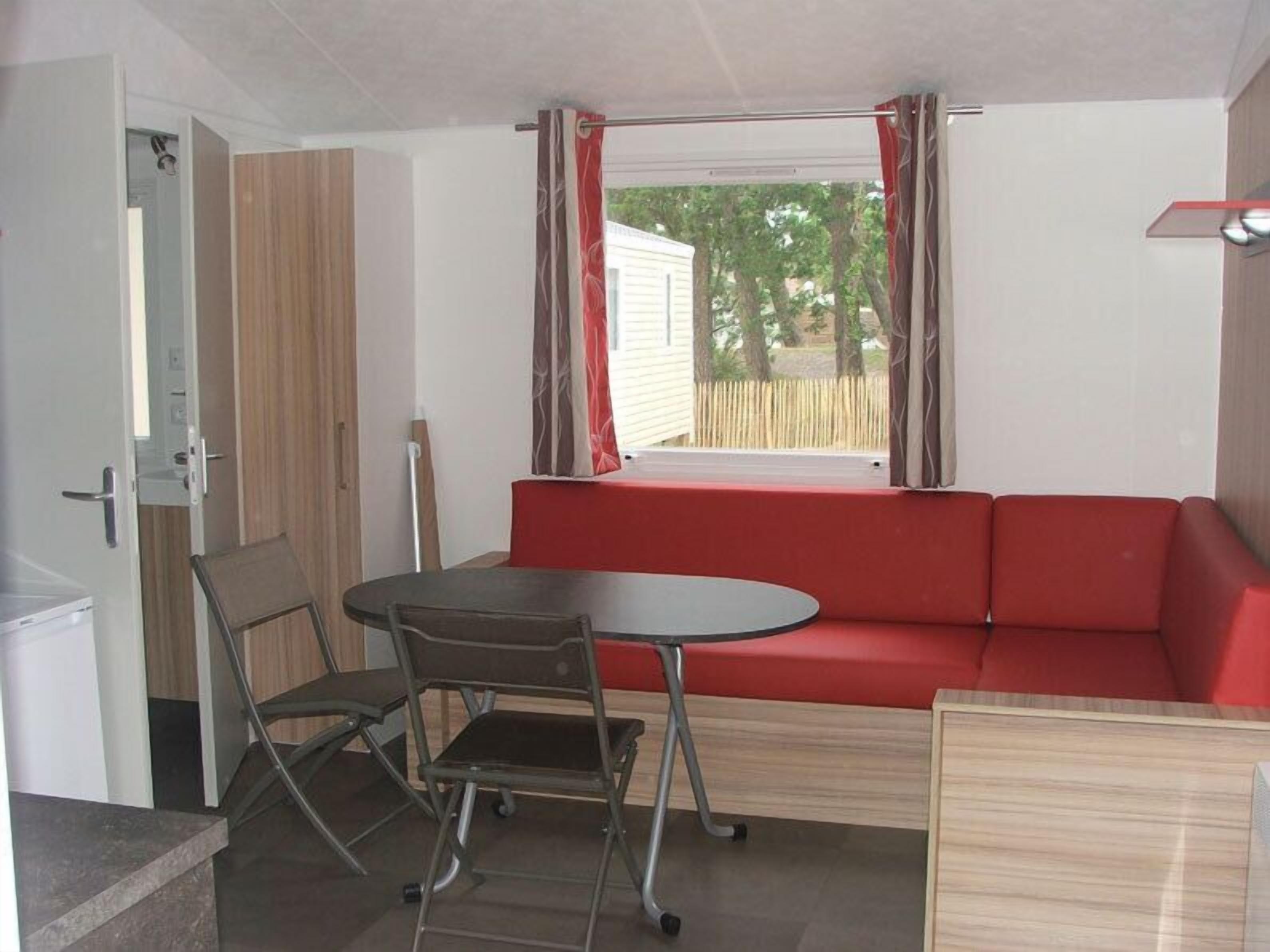 A compact room with a red sofa, a small round table, and a folding chair. A window with curtains is visible, and a door is open to the left.