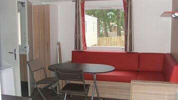 A compact room with a red sofa, a small round table, and a folding chair. A window with curtains is visible, and a door is open to the left.