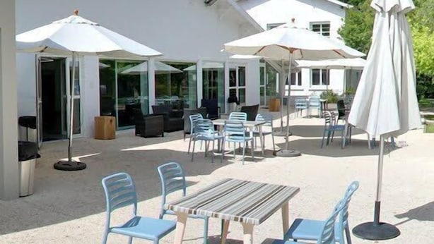 An outdoor patio area with tables and chairs, umbrellas, and a building with large windows.