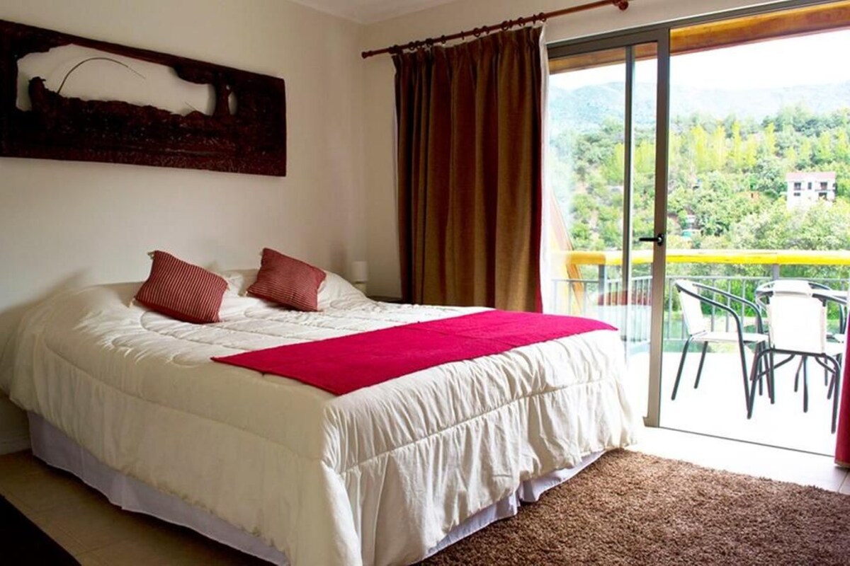 A bedroom with a large bed, a red throw blanket, and a view of a balcony with chairs.