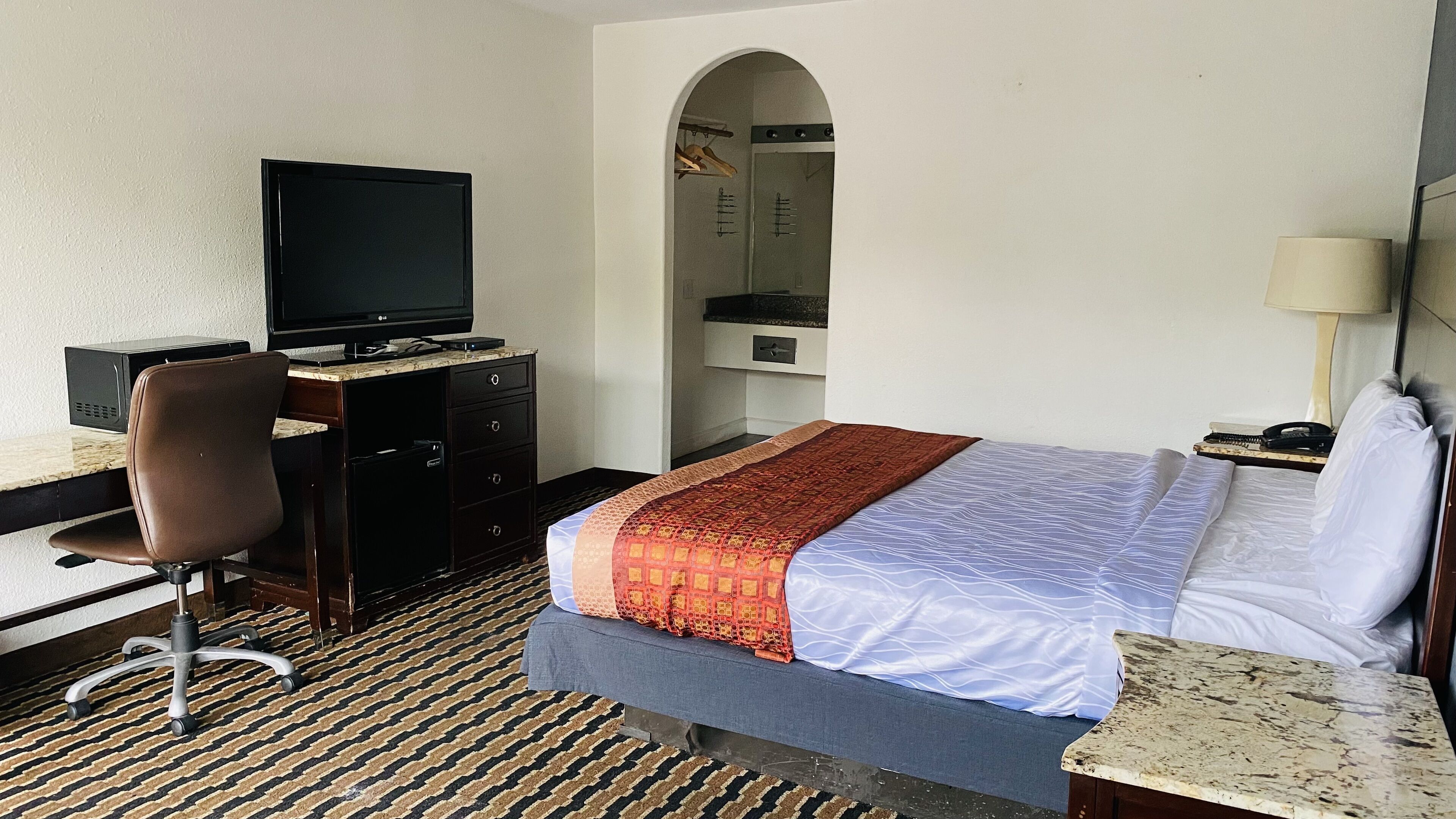 A hotel room with a bed, a desk with a television, a chair, a desk lamp, and a small table.