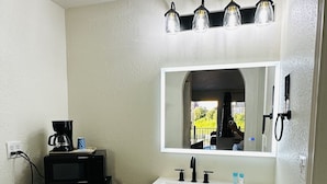 Un baño con un lavabo blanco, un espejo y una cafetera negra sobre una mesita.