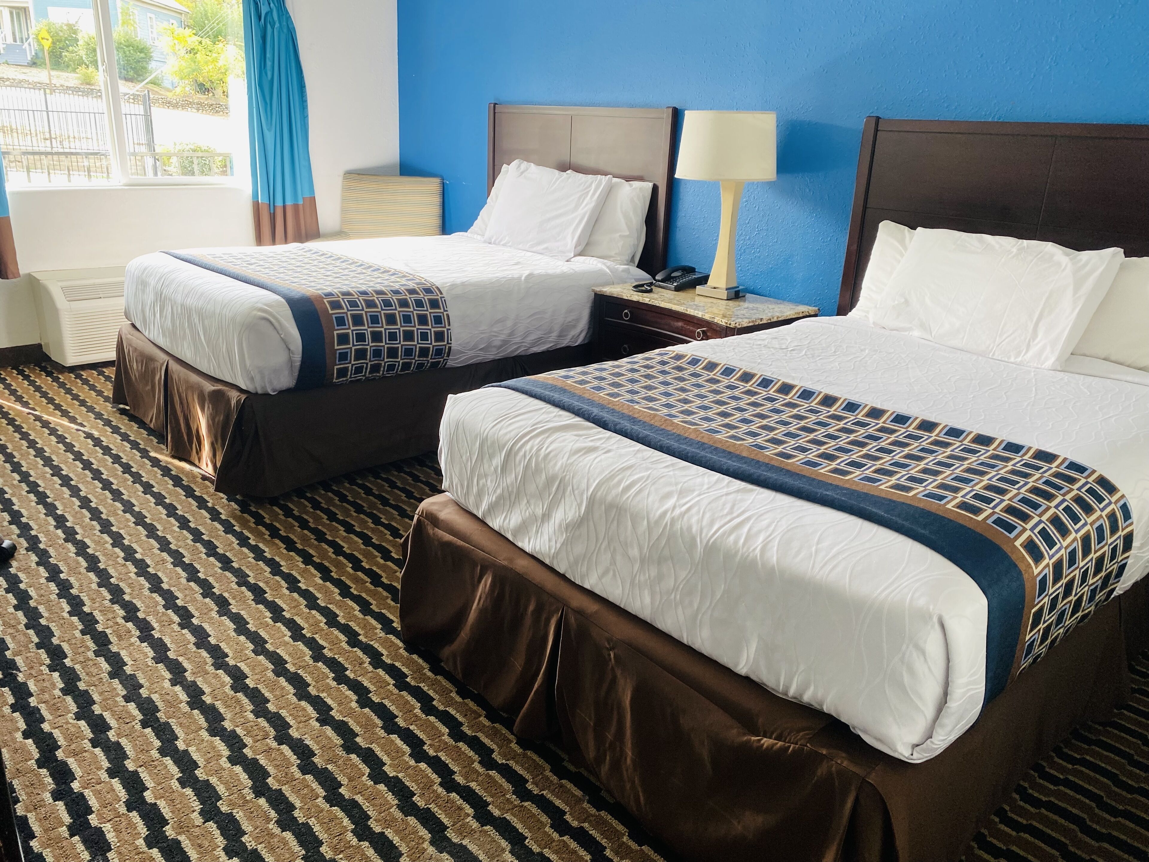 A hotel room with two beds, a patterned carpet, and a view of the outside.