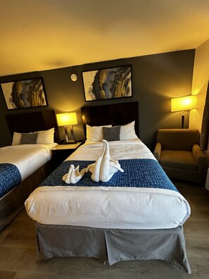 A hotel room with two beds, a large bed with a blue and white patterned bedspread and two swan-shaped pillows, and a smaller bed with a plain white bedspread. There are two wall-mounted lamps, a chair, and two framed pictures on the wall. - Sierra Inn (San Andreas)