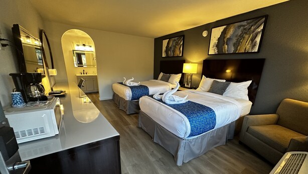 Habitación de hotel con dos camas, un escritorio con cafetera, un sofá y obras de arte en las paredes.