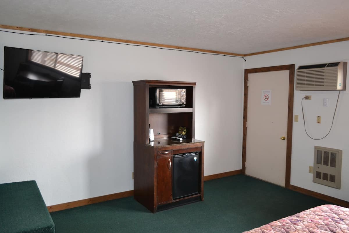 A small room with a wooden cabinet containing a microwave, a small refrigerator, and a television mounted on the wall. There is a door with an air conditioning unit on the wall.