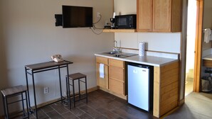 A compact kitchen with a microwave, coffee maker, and a small refrigerator. There is a wooden cabinet above the sink and a wooden table with a chair. - The Village Inn Motel and Restaurant (Challis)