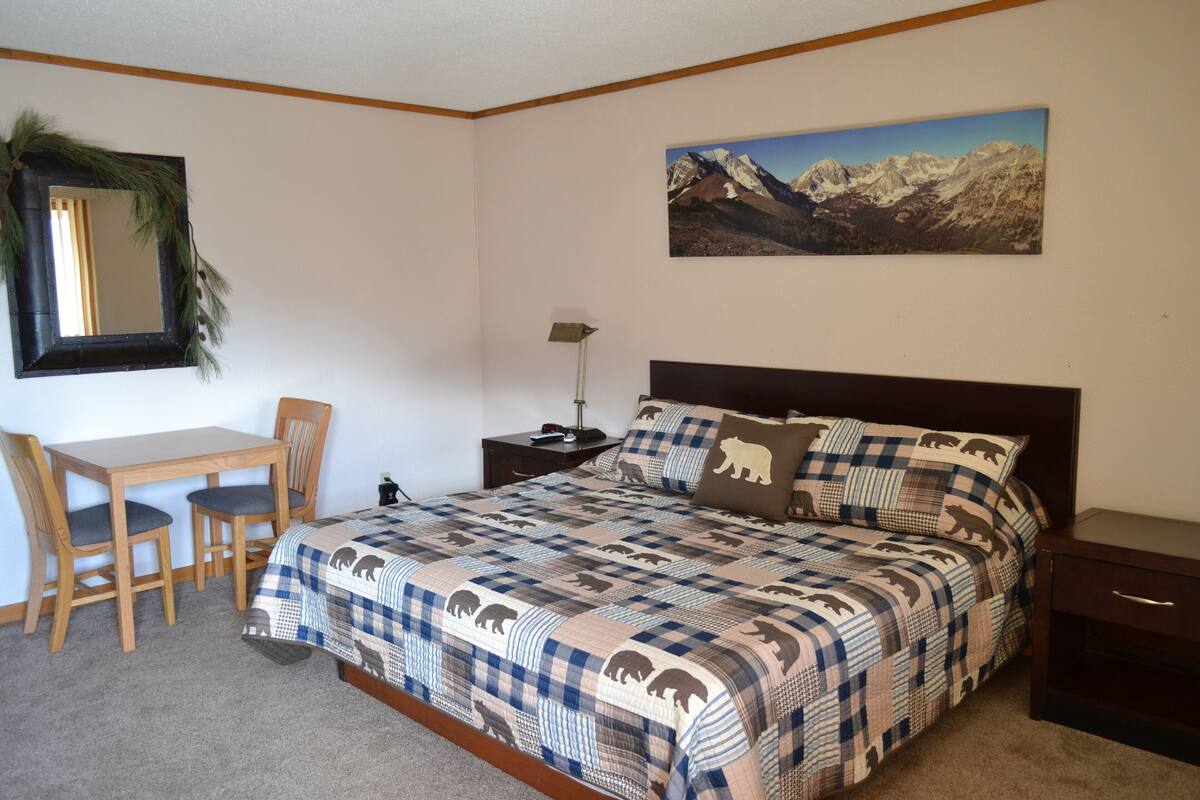 A bedroom with a bed, a desk, two chairs, a nightstand, a lamp, and a mountain landscape painting on the wall.