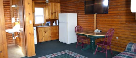 A wooden cabin interior with a kitchen, dining area, and a living room with a plaid sofa.