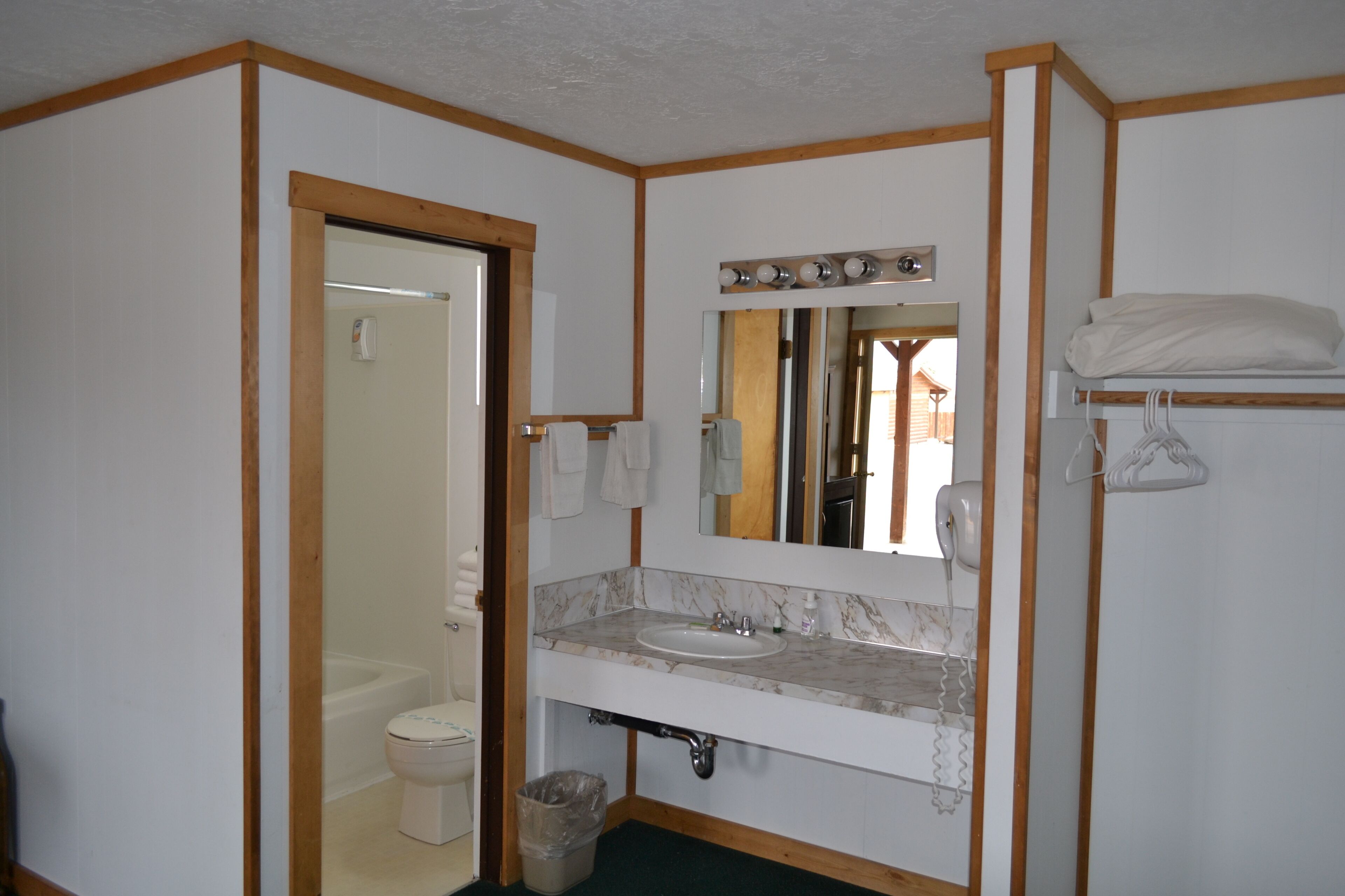 A bathroom with a white sink, mirror, and wooden accents. There is a toilet and a trash can visible.