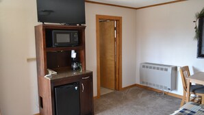 A compact room with a kitchenette area including a microwave, a small refrigerator, and a coffee maker. There's a television on a wooden stand above the microwave. A door leads to another room, and a radiator is mounted on the wall. - The Village Inn Motel and Restaurant (Challis)