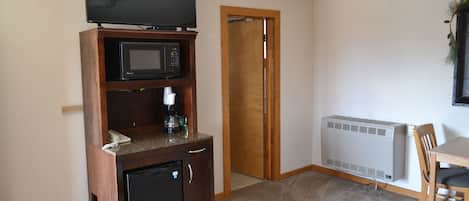 Habitación funcional con kitchenette que incluye microondas, refrigerador pequeño y cafetera. Hay un televisor sobre un soporte de madera encima del microondas. Una puerta conduce a otra habitación y un radiador está montado en la pared.