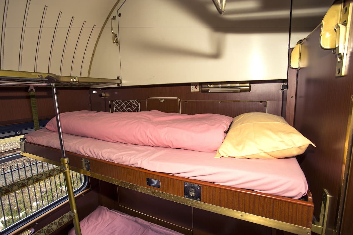 A train compartment with a bunk bed, a window with metal bars, and overhead luggage racks.