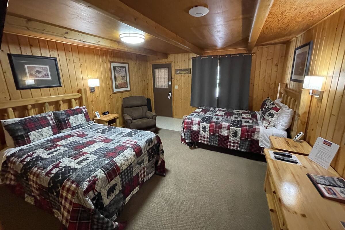A wooden cabin room with two beds, a chair, a desk, and a nightstand.