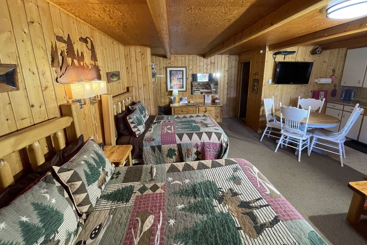 A cozy cabin-style room with wooden walls and ceiling, a bed with a quilt, a desk with a chair, and a television.