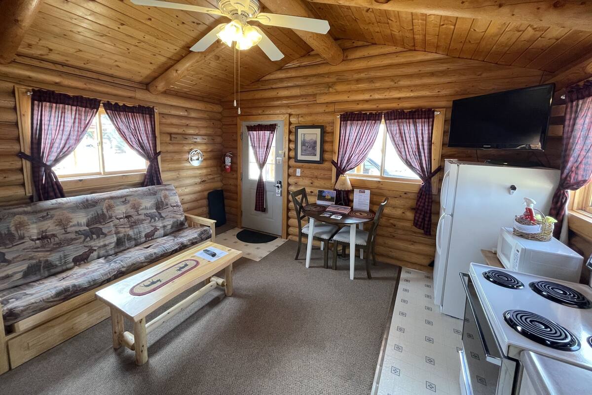 A log cabin interior with a sofa, dining table, chairs, a ceiling fan, a television, and a kitchen area with a stove.