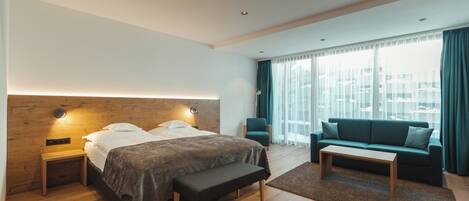 A modern hotel room with a wooden headboard bed, a bench, a sofa, and a large window with curtains.
