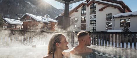 Un couple se détend dans un bain à remous, la vapeur s'élevant autour d'eux, entouré d'une terrasse en bois et d'un bùtiment au toit en pente, le tout dans un paysage enneigé.