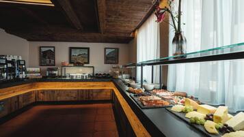 Ein Bereich mit Buffet, der eine Auswahl an Käse, Fleisch und Früchten bietet. Eine Kaffeestation mit mehreren Kaffeemaschinen und einer Präsentation von Kaffeetassen. Der Raum verfügt über Holzbalken an der Decke und ein großes Fenster mit transparenten Vorhängen.