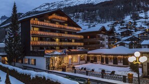A multi-story hotel with balconies adorned with string lights, a stone facade, and a view of snow-covered mountains. - Zermatt Budget Rooms at Alpen Resort, Spa & Dining (Zermatt)