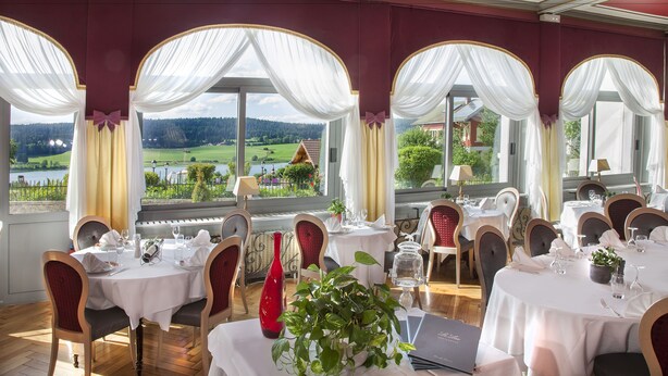 A dining area with tables set for a meal, large windows with curtains, and a view of a green landscape. - Hotel beau site (Malbuisson)
