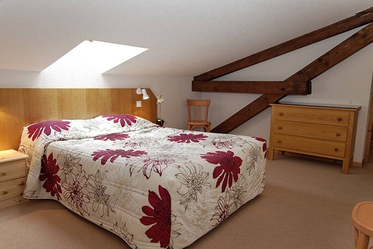 A bedroom with a bed, a nightstand, a wooden dresser, a chair, and a sloped ceiling with exposed beams.