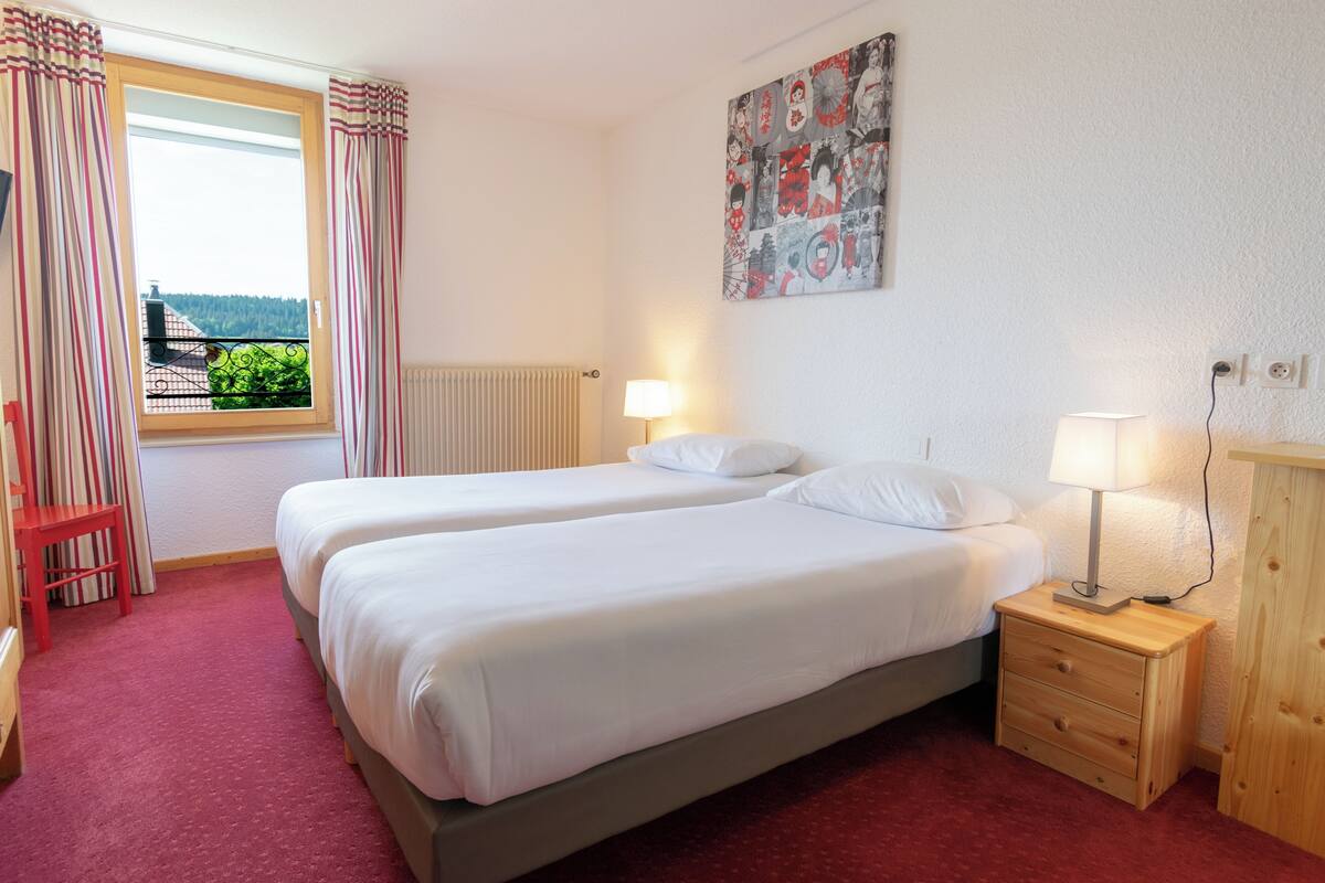 A hotel room with two single beds, a wooden nightstand with a lamp, a red chair, a window with curtains, and a wall with a poster.
