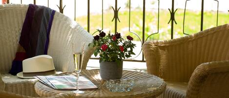 Una silla de mimbre con un sombrero blanco y una copa de vino sobre una mesa, una planta en maceta con flores rojas, y una vista de follaje a través de una barandilla de metal.