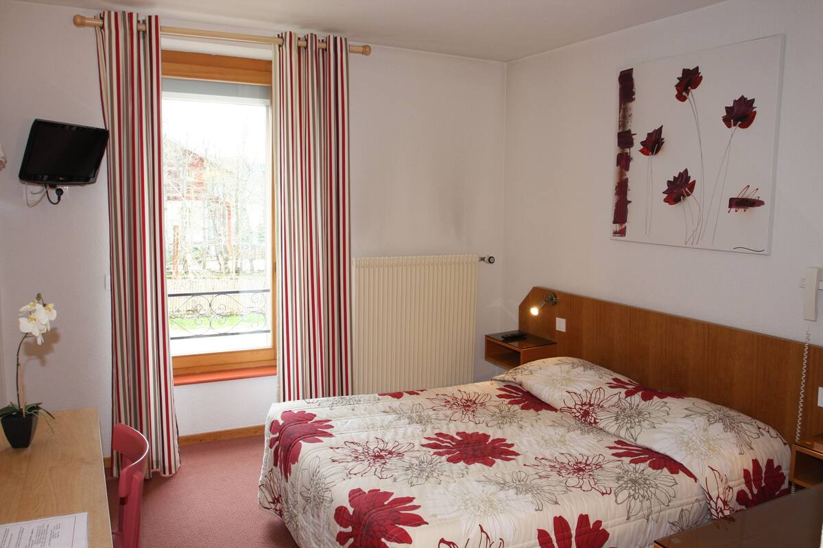A bedroom with a bed, a desk, a chair, a TV, and a window with curtains.
