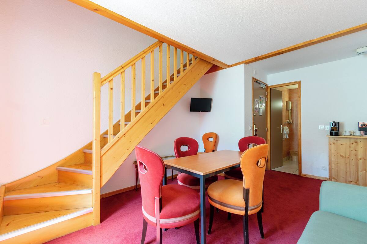A room with a wooden staircase, a dining table with chairs, a television mounted on the wall, and a wooden cabinet.