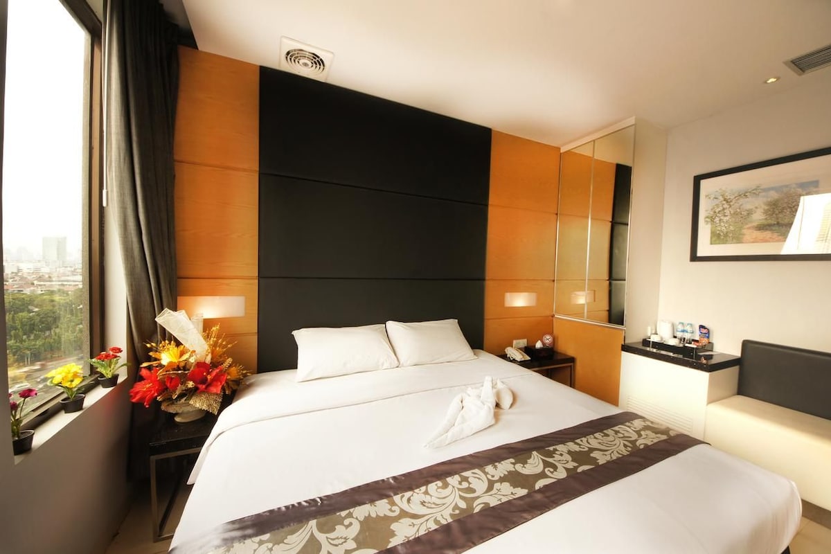 A hotel room with a large bed, a nightstand with a lamp, a window with curtains, and a view of the city.