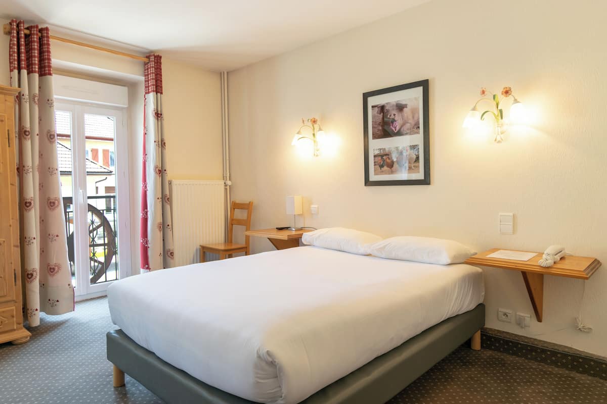 A hotel room with a large bed, a desk with a chair, a small table with a lamp, and a wall-mounted phone.