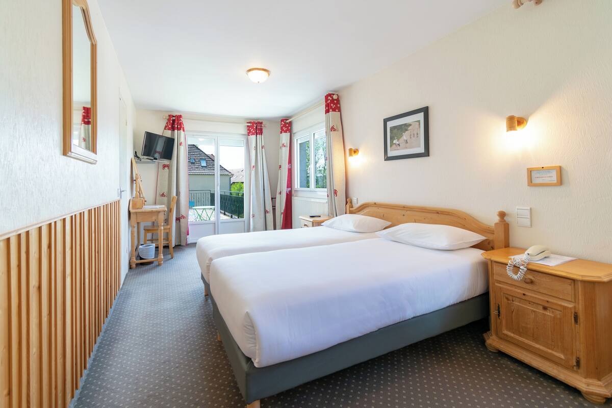 A hotel room with two beds, a wooden nightstand, a desk with a chair, a television, and a window with curtains.
