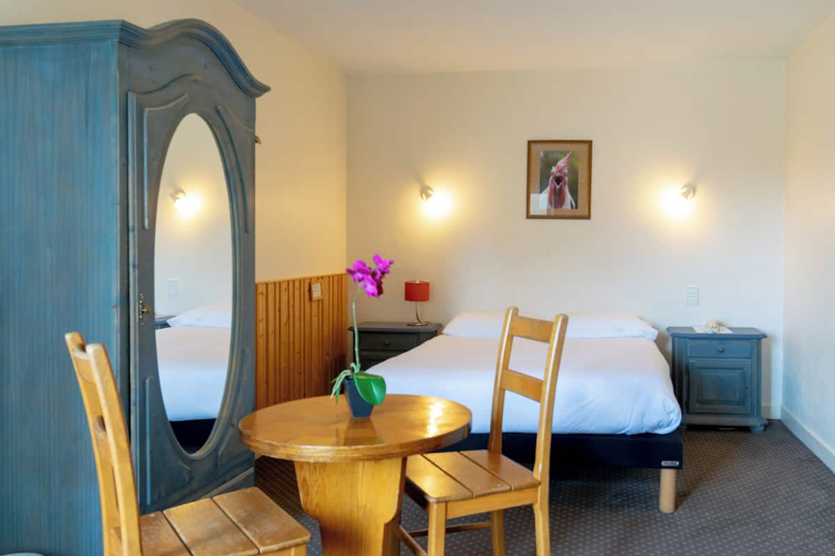 A hotel room with a bed, a wooden chair, a round table with a vase of flowers, a dresser, and a nightstand.