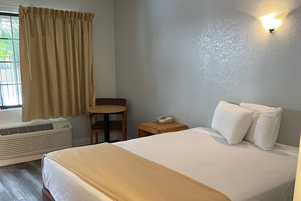 A hotel room with a bed, a bedside table with a phone, a chair, and a window with curtains.