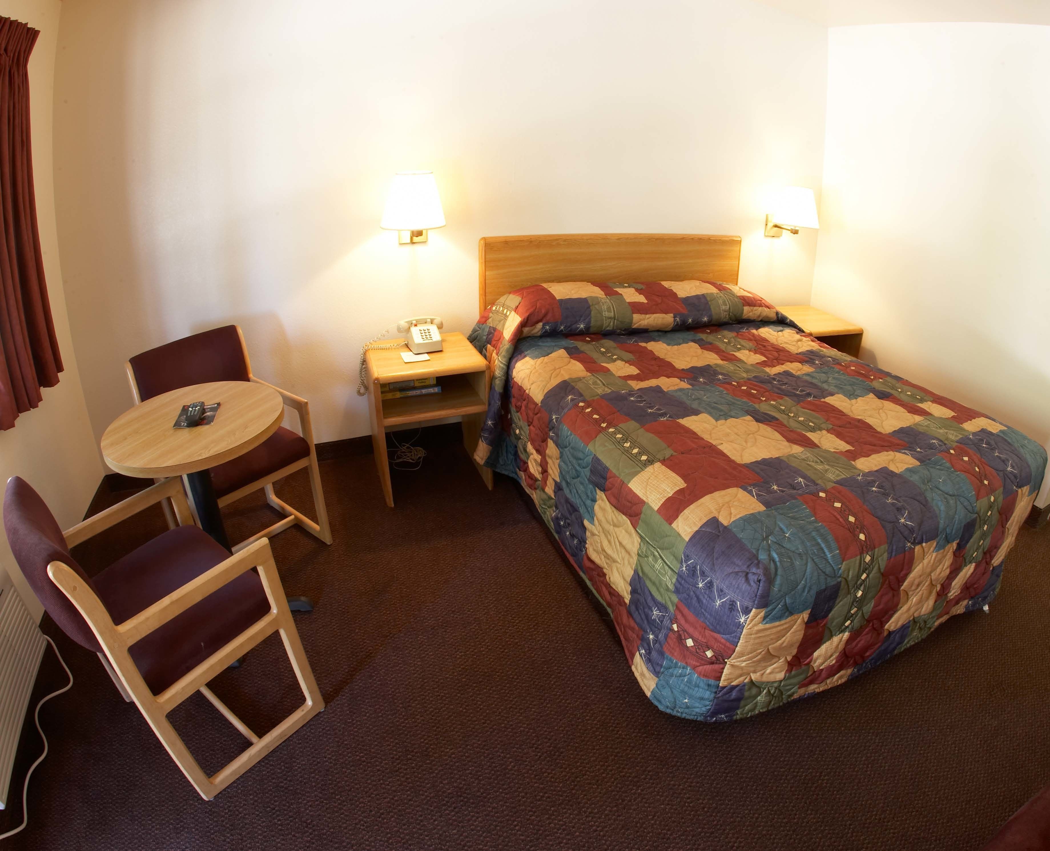A hotel room with a bed, a small table with a chair, a bedside table with a lamp, and a telephone.