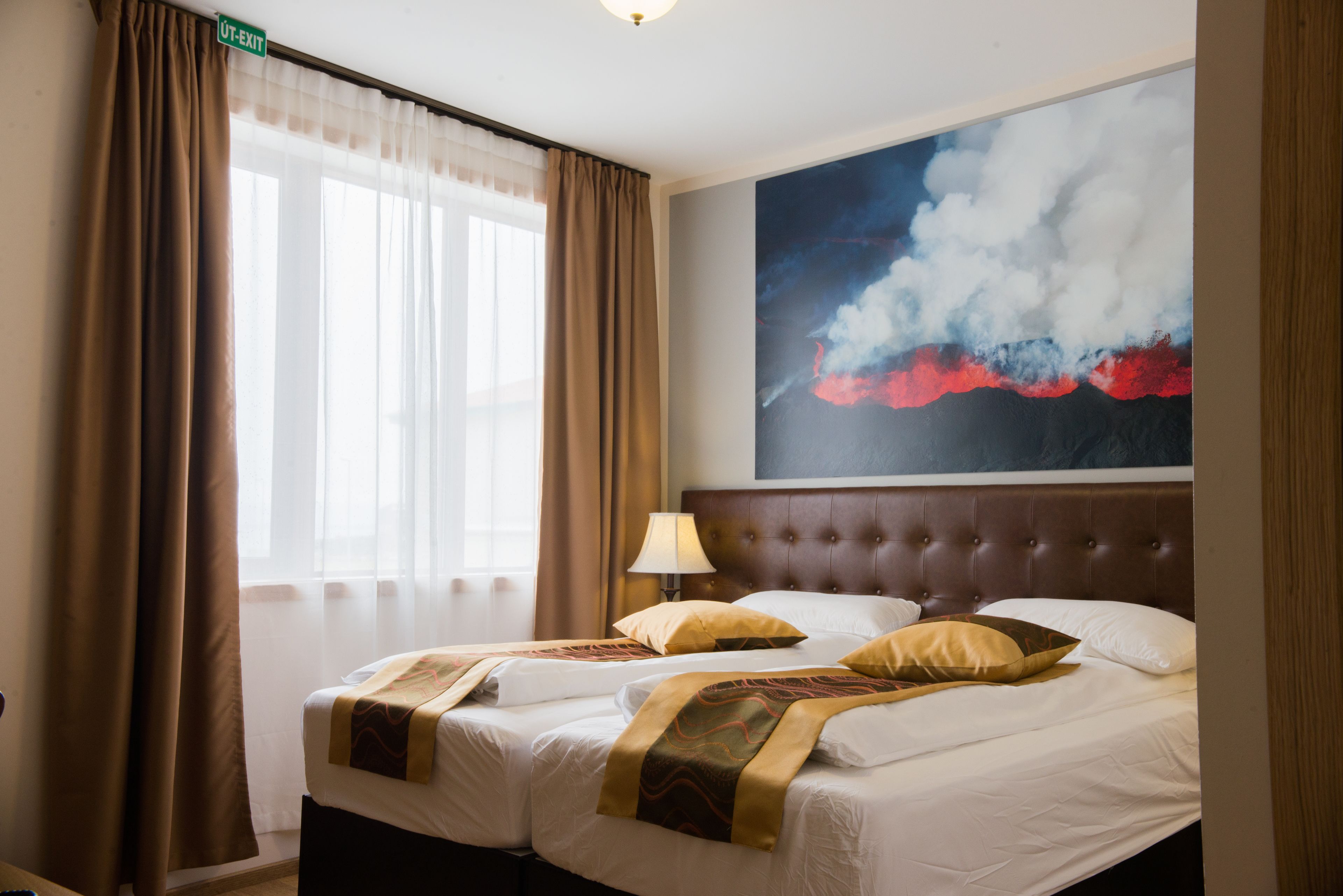 A hotel room with a large bed, a headboard, a window with curtains, and a painting on the wall.