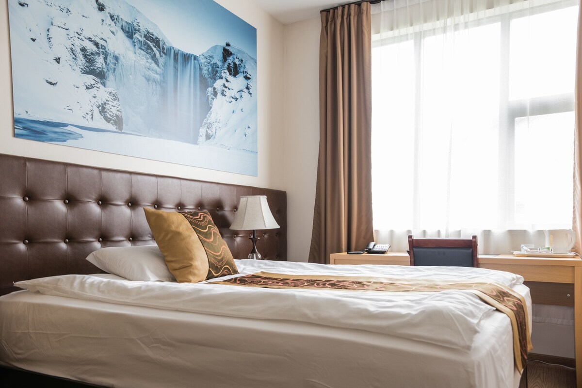 A hotel room with a large bed, a headboard, a desk, a chair, a lamp, a painting of a waterfall, and a window with curtains.