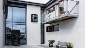 Entrada de un hotel con puerta de cristal, un cartel que dice "Hotel Jazz", un balcón con plantas en macetas y un banco en el exterior.