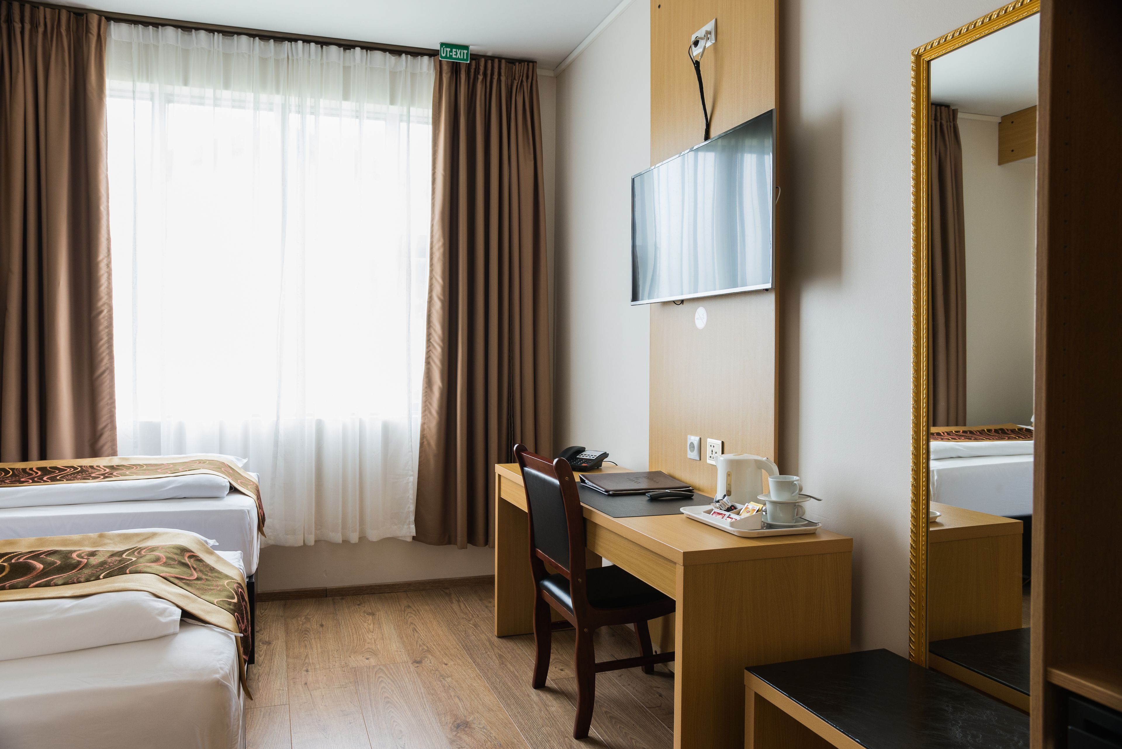 A hotel room with two beds, a desk, a chair, a TV, a mirror, and a window with curtains.