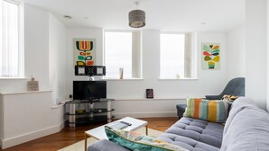 A modern living room with a grey sofa, a white coffee table, and a flat-screen TV. There are two windows with blinds, a ceiling light, and artwork on the walls. - Birmingham Serviced Apartments Hagley Rd (Birmingham)