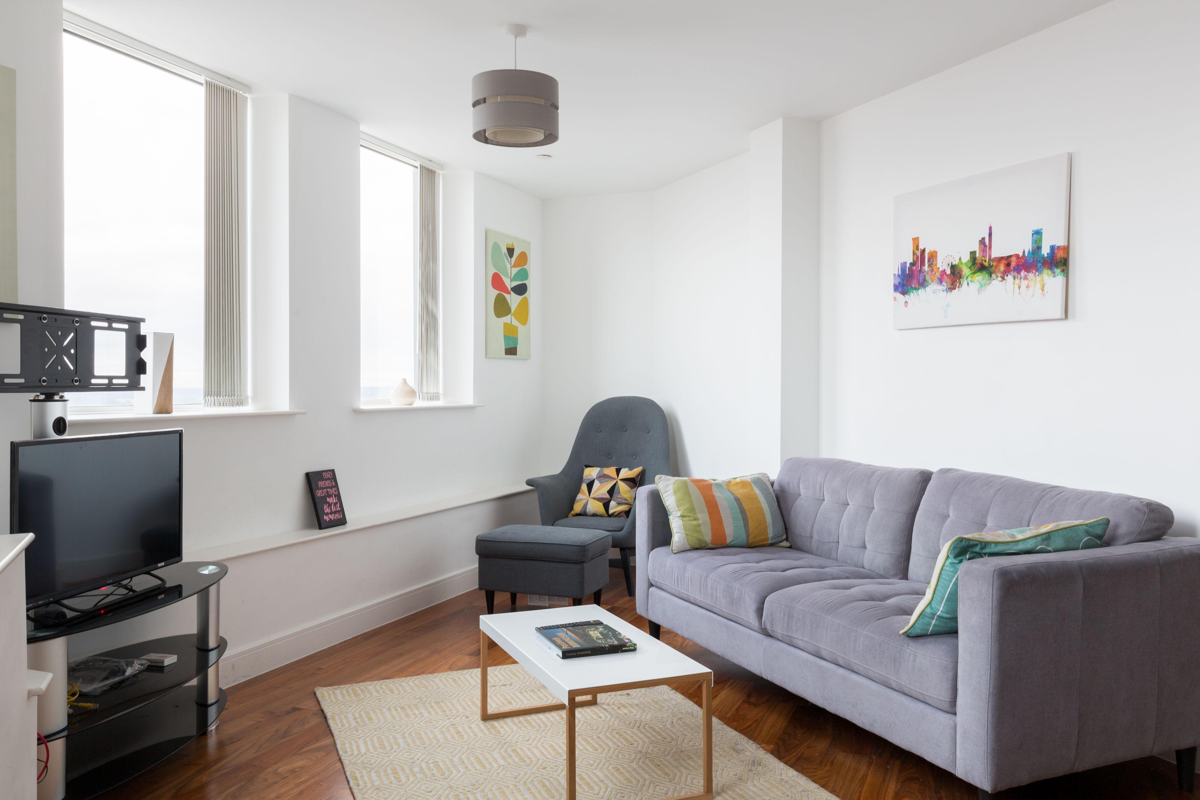 Ein modernes Wohnzimmer mit einem grauen Sofa, einem kleinen Tisch, einem Fernseher und einem Stadtbild an der Wand.