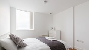 A minimalist bedroom with a bed, a window, a bedside table, and a wall-mounted heater. - Birmingham Serviced Apartments Hagley Rd (Birmingham)