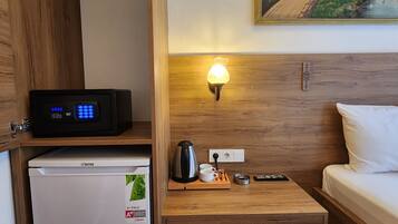 Quarto de hotel pequeno com uma cama, uma mesinha com abajur, um refrigerador, uma cafeteira e um quadro na parede.