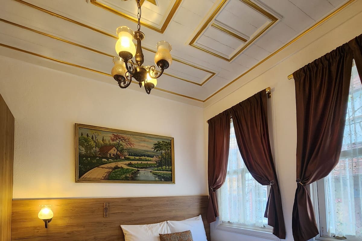 A hotel room with a large bed, a nightstand with a lamp, a painting on the wall, and a window with curtains.