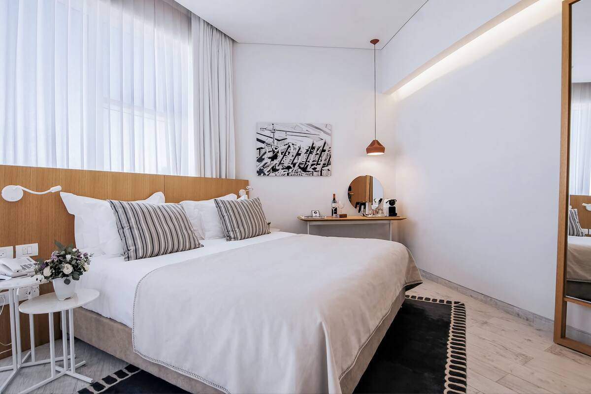 A modern hotel room with a large bed, bedside tables, a mirror, and a cityscape artwork on the wall.